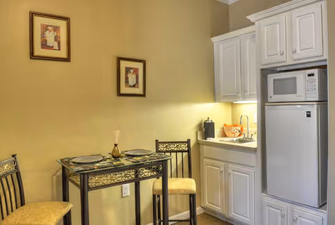 Small dining area with a glass-top table set for two, two chairs with cushioned seats, and a kitchenette featuring white cabinets, a microwave, a small refrigerator, and a sink. Two framed pictures hang on the beige wall above the table.