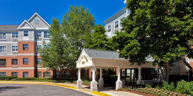 Exterior view of Pin Oak Village senior living facility showing a multi-story building with a covered entrance, surrounded by trees and landscaping under a clear blue sky.