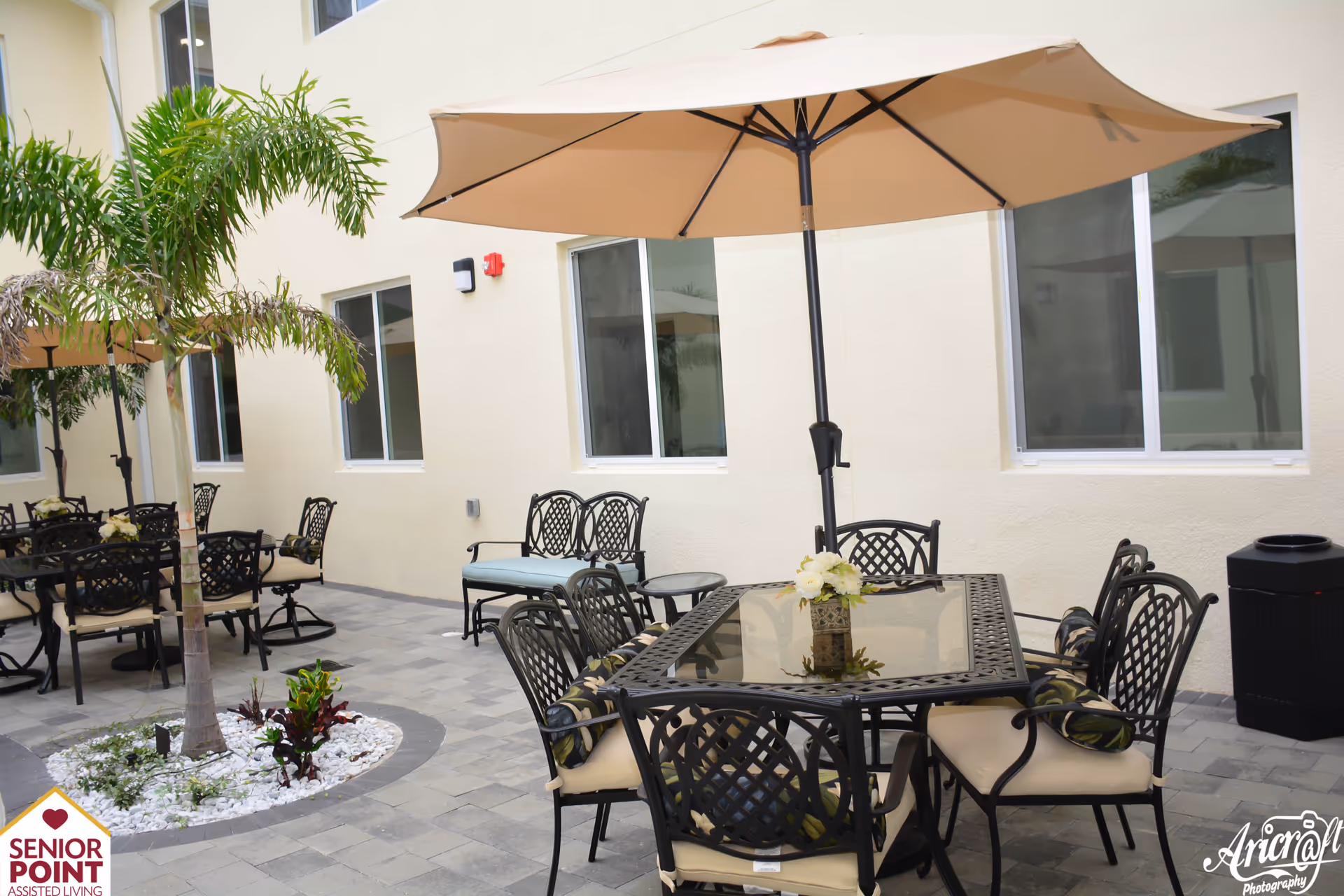Outdoor patio area at Senior Point Assisted Living featuring multiple black metal tables with glass tops and matching chairs with beige cushions. Each table has a large beige umbrella providing shade. There are small landscaped areas with palm trees and plants surrounded by white stones. The patio is paved with gray tiles and is adjacent to a cream-colored building with several windows.