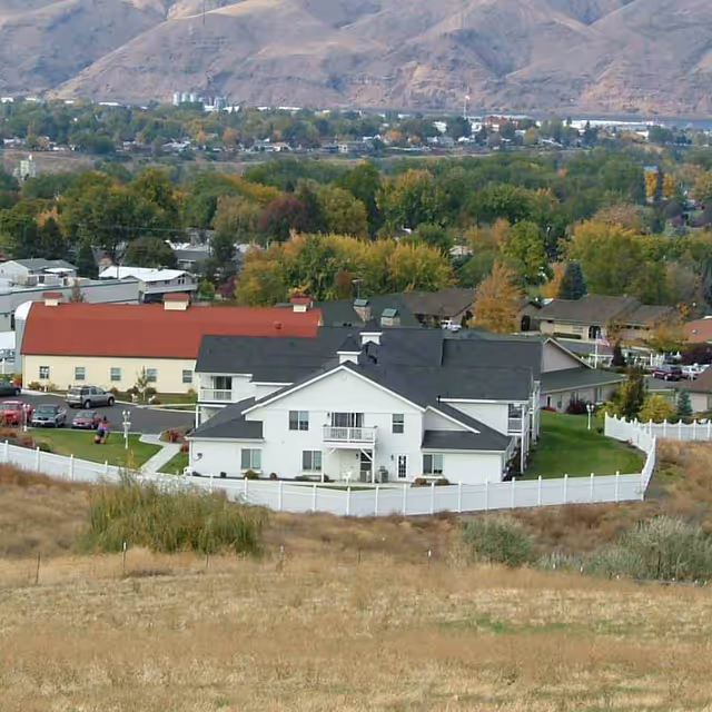 Aerial view of a senior living facility named Generations at Lewiston, featuring multiple buildings with white and red roofs, surrounded by a white fence. The facility is set against a backdrop of trees with autumn foliage and distant mountains.