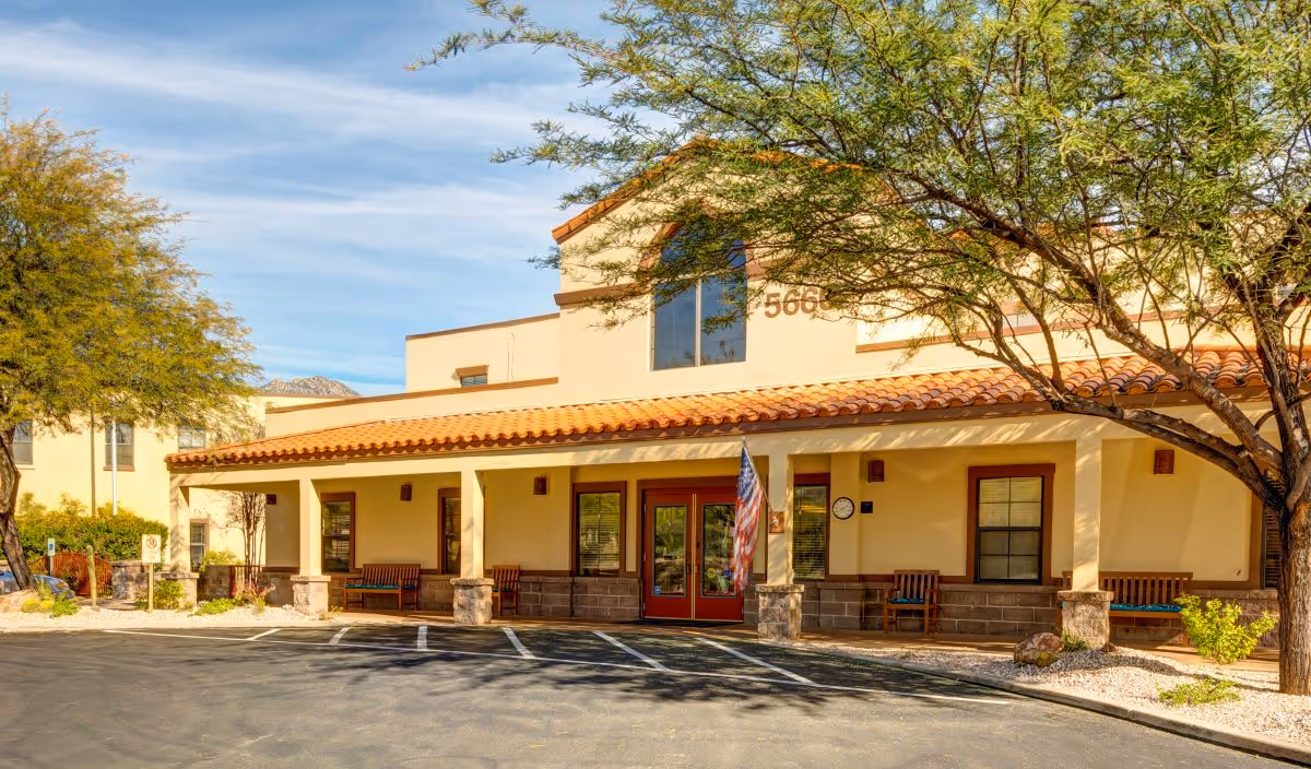 Exterior view of a senior living facility building with a tiled roof, beige walls, and a covered entrance supported by columns. There are benches along the front porch, an American flag near the entrance, and trees providing shade. The sky is clear with some clouds.