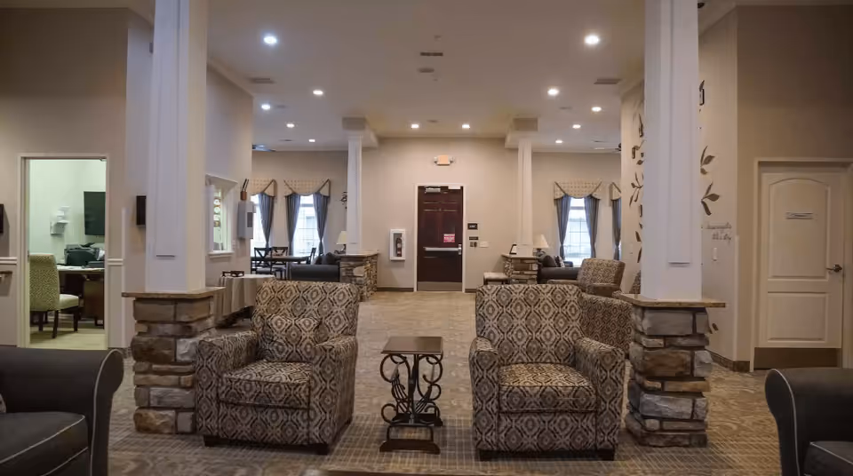 A spacious and well-lit common area in a senior living facility featuring patterned armchairs, a small decorative table, stone pillars, and windows with curtains. The room has a neutral color palette with beige walls and carpeted floors, and there are additional seating and tables visible in the background.