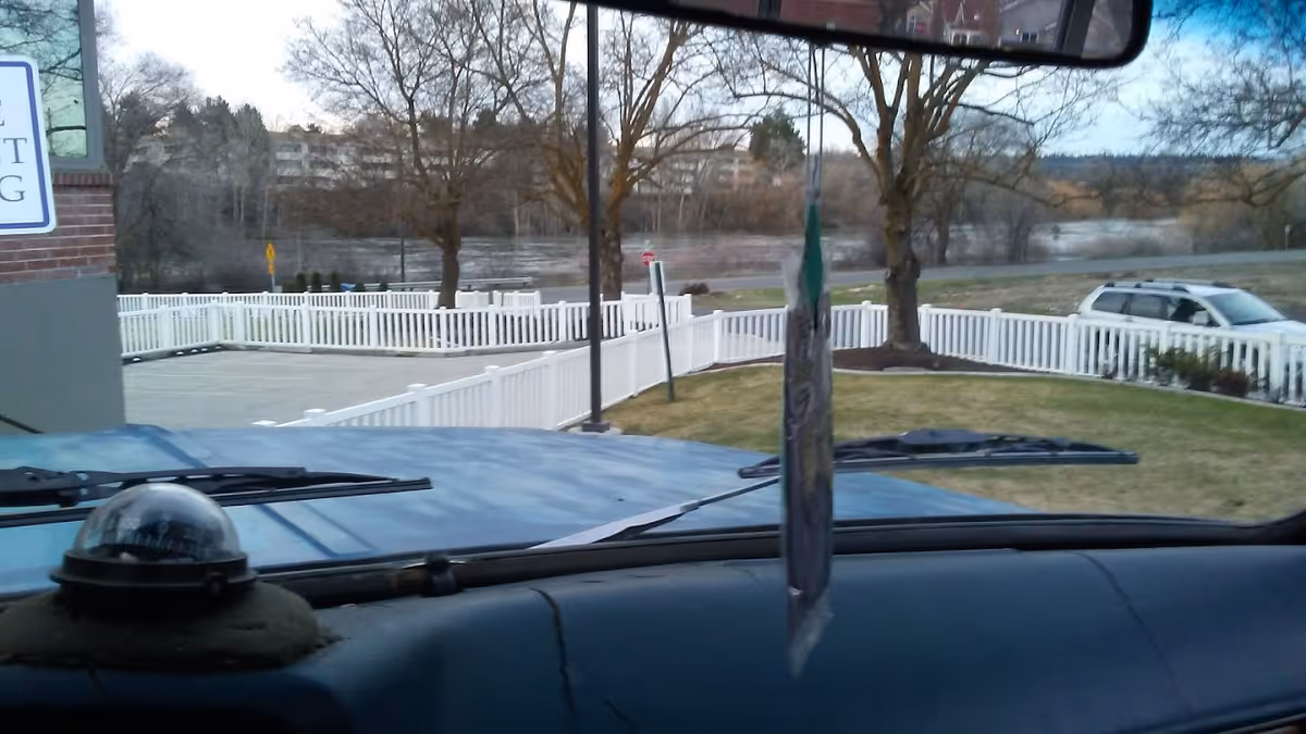 View from inside a vehicle looking out through the windshield at a fenced grassy area with trees and a river in the background. A white fence encloses the area, and a parked white SUV is visible on the right side. The dashboard of the vehicle and a hanging air freshener are also visible.