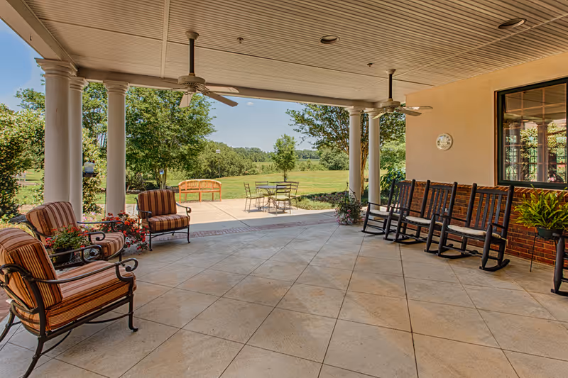 Covered outdoor patio area with ceiling fans, several cushioned chairs, rocking chairs, and a small table with chairs. The patio overlooks a green lawn with trees and a bench in the distance under a clear blue sky.