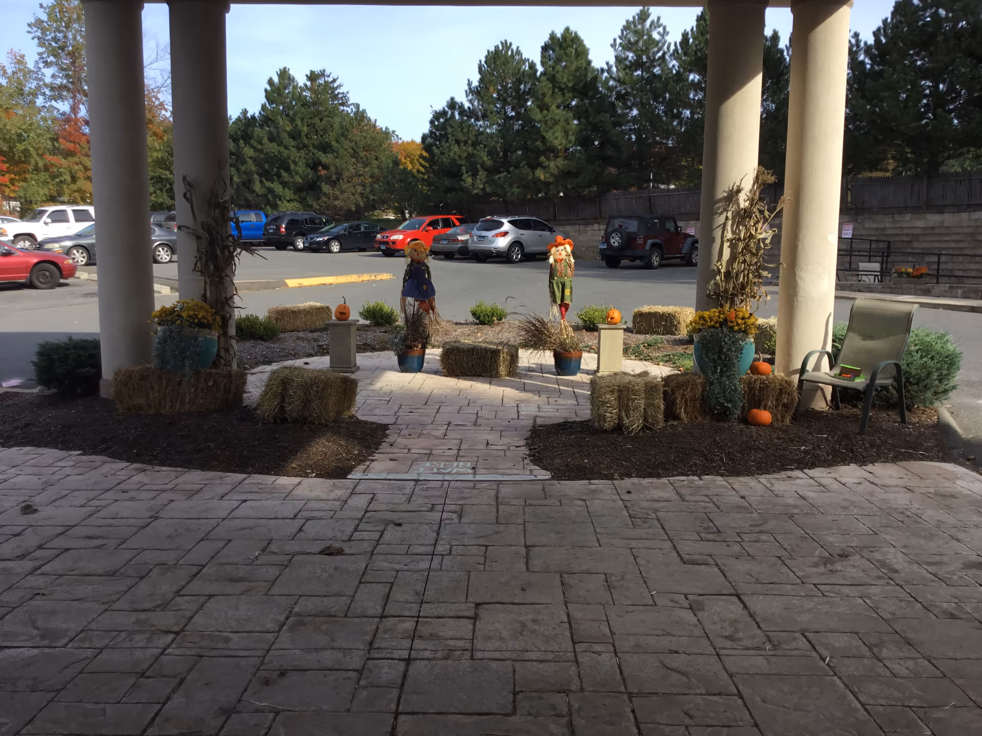 Covered entrance to a care facility with columns, autumn decorations (hay bales, pumpkins, scarecrows) and a view of the parking lot.