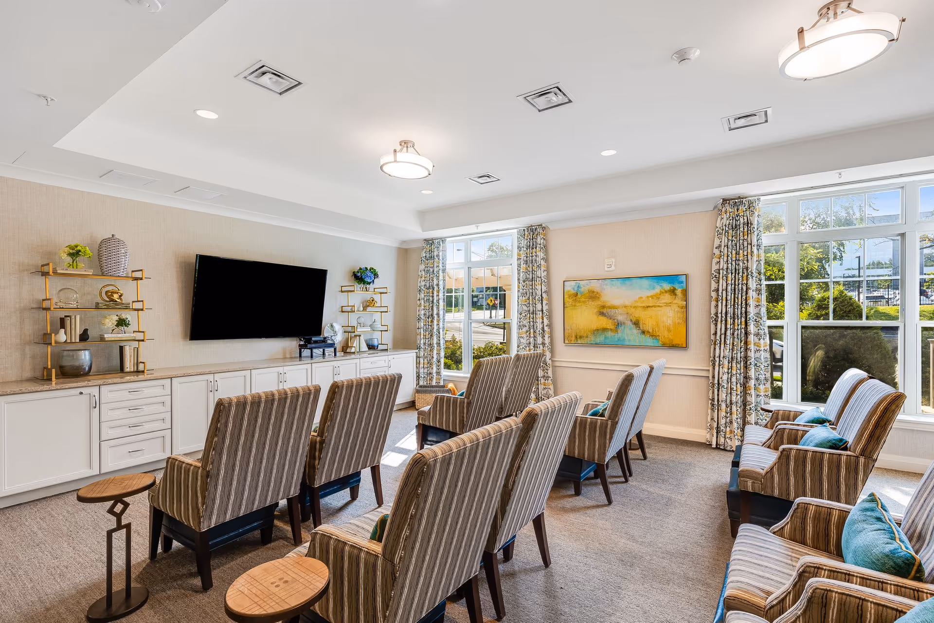 Bright common living room with rows of striped armchairs facing a wall-mounted TV, large windows, and decorative shelving.