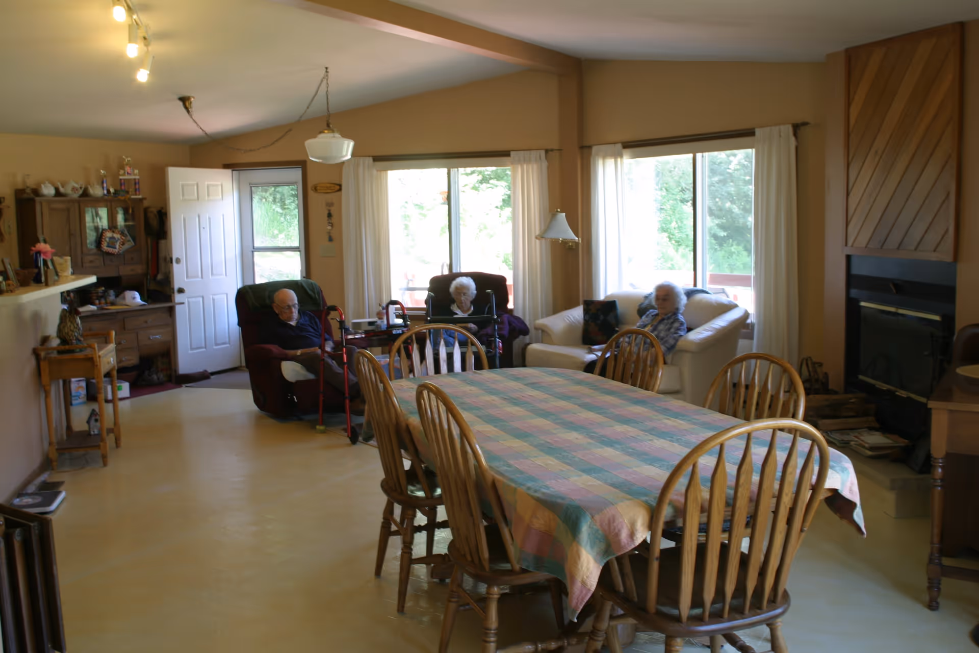 An open-plan dining and living room with a long table and chairs in the foreground and elderly residents seated on sofas and recliners near windows and a fireplace.