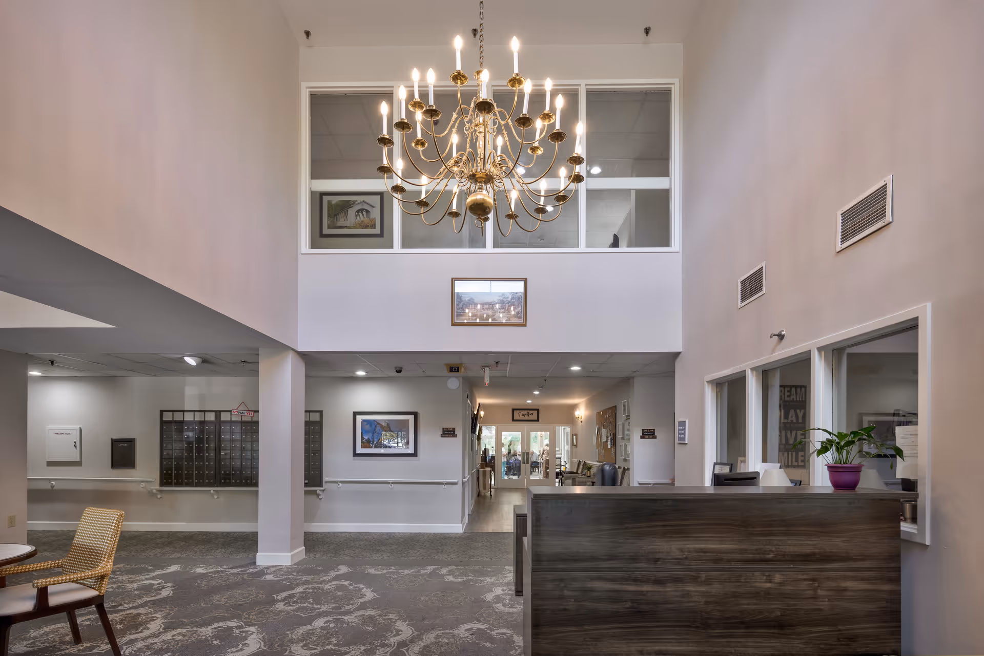 Spacious two-story lobby with a large chandelier, reception desk, mailboxes, seating, and a hallway leading further into the facility.