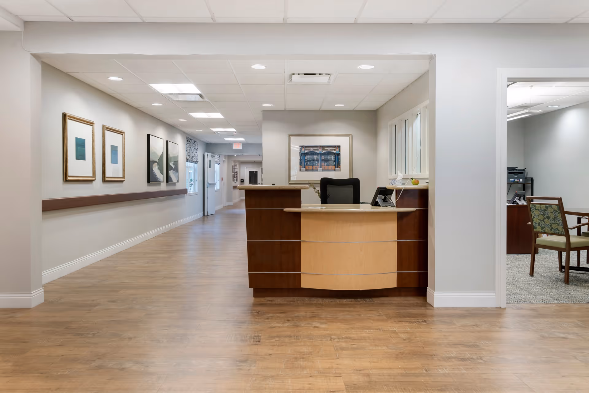 Reception desk area in a senior living facility with a wooden desk, black office chair, and framed artwork on the walls. The hallway has wooden flooring, light-colored walls, and ceiling lights. To the right, there is an open doorway leading to a room with chairs and office equipment.