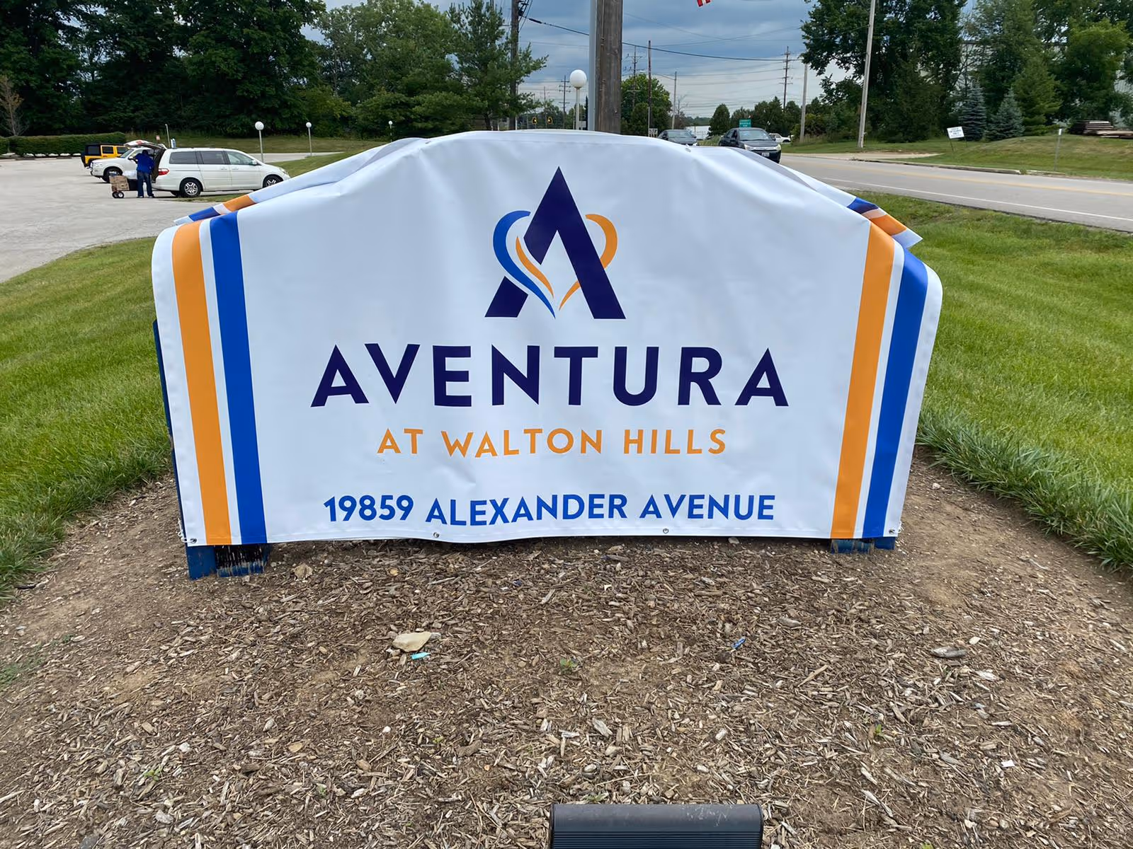 Outdoor banner sign on a lawn reading "Aventura at Walton Hills" with the address and a road and parked cars in the background.
