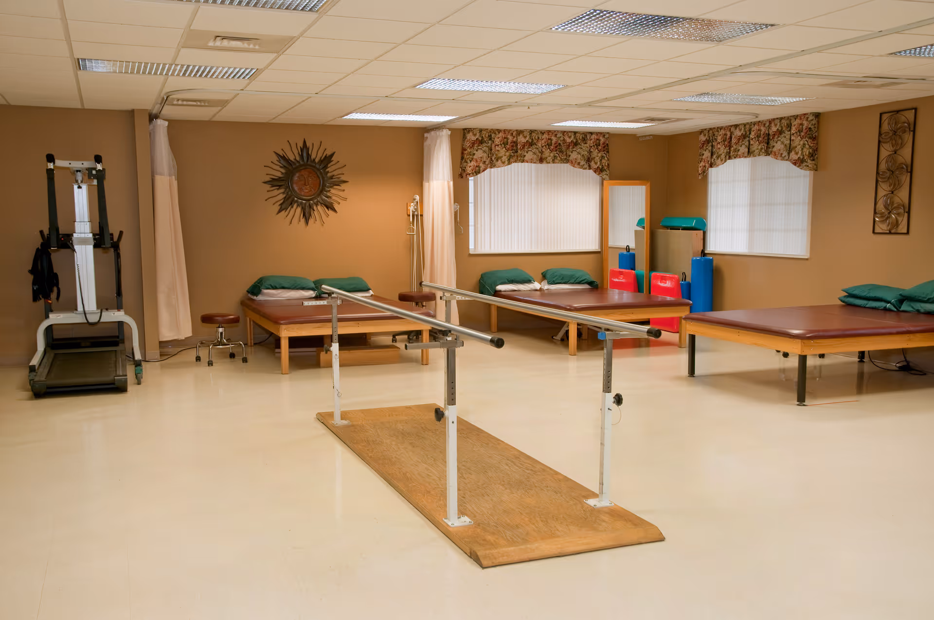 A physical therapy room with three padded therapy tables, a set of parallel bars for walking exercises, a treadmill, and various therapy equipment. The room has beige walls, two windows with floral valances, and a sun-shaped wall decoration.