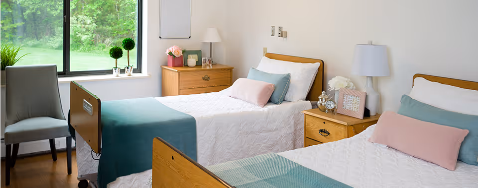 A bright and clean bedroom with two single beds, each made with white quilts, teal and pink pillows, and teal blankets. There are two wooden nightstands with lamps, flowers, and decorative items between and beside the beds. A gray chair is placed near a window showing green outdoor foliage.