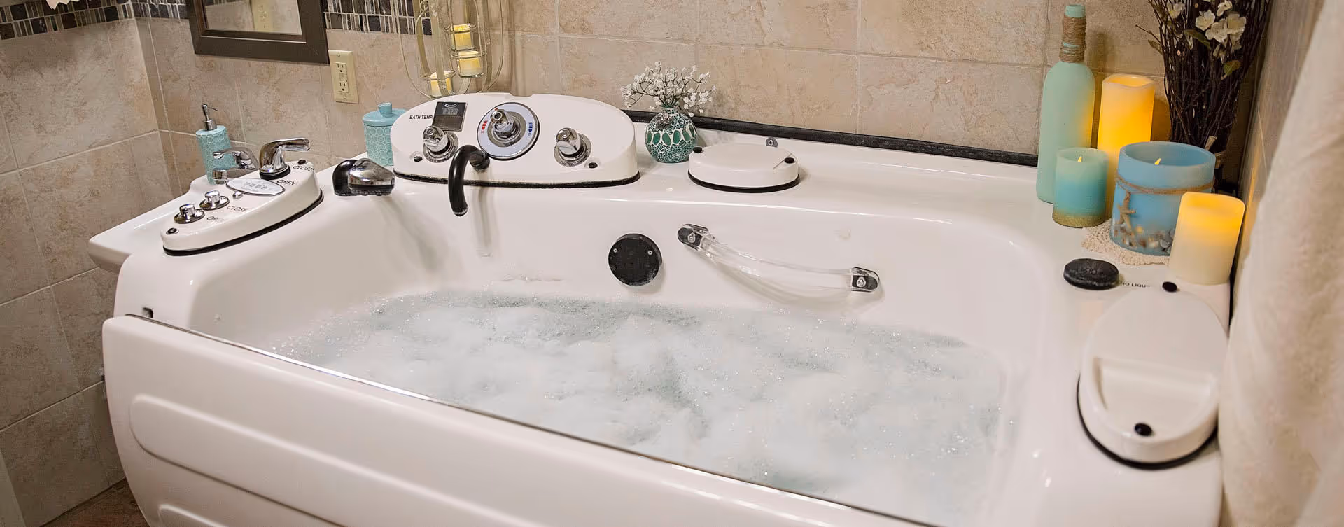 A white whirlpool bathtub filled with bubbly water in a tiled bathroom. The tub has multiple control knobs and a faucet, with decorative candles and a small plant placed on the edges. The walls are tiled in beige tones.