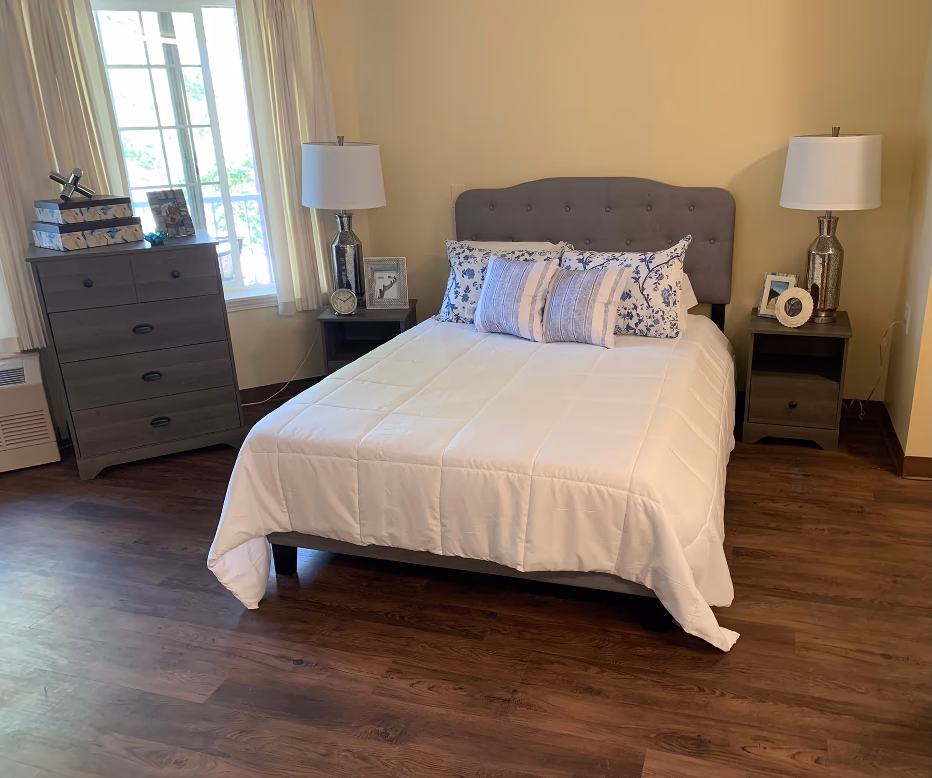 A neatly made bed with a gray upholstered headboard, white comforter, and decorative pillows in a bedroom with wooden flooring. There are two matching nightstands on either side of the bed, each with a silver lamp and framed photos. A chest of drawers with decorative items is positioned near a window with light-colored curtains.