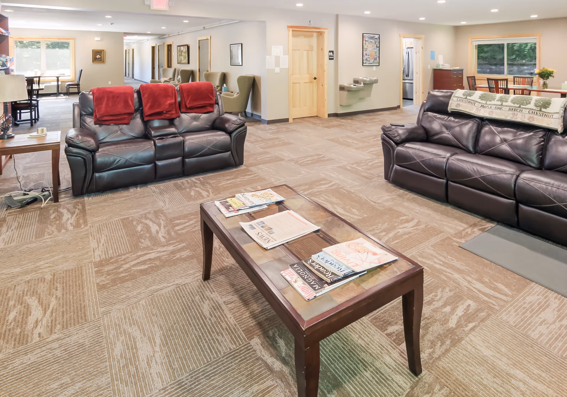 A spacious living room area in South Torch Assisted Living featuring two dark leather sofas with red and patterned throws, a wooden coffee table with magazines, beige patterned carpet, and a background showing a hallway with armchairs, a dining area with tables and chairs, and a water fountain near a wooden door.