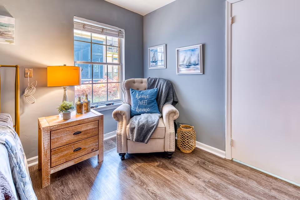 Cozy bedroom corner with an upholstered armchair draped with a throw and pillow, a wooden nightstand with a lamp, and a window.