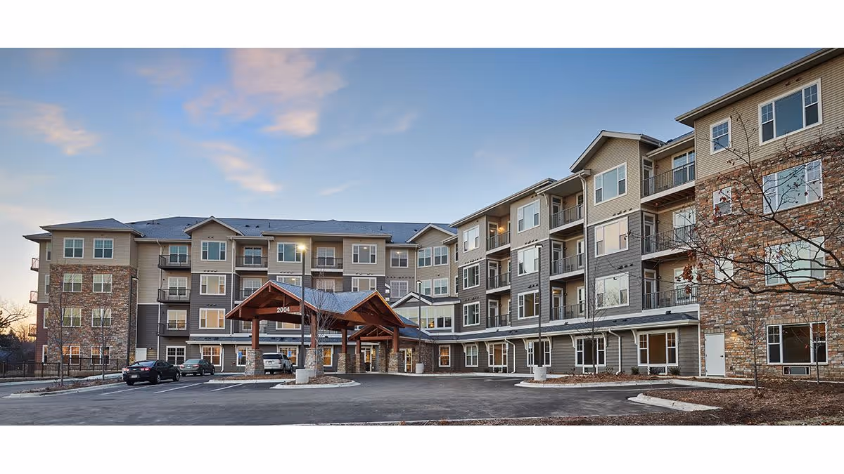 Exterior view of a multi-story senior living facility building with a covered entrance and several balconies, surrounded by a parking area and some trees under a partly cloudy sky during dusk.