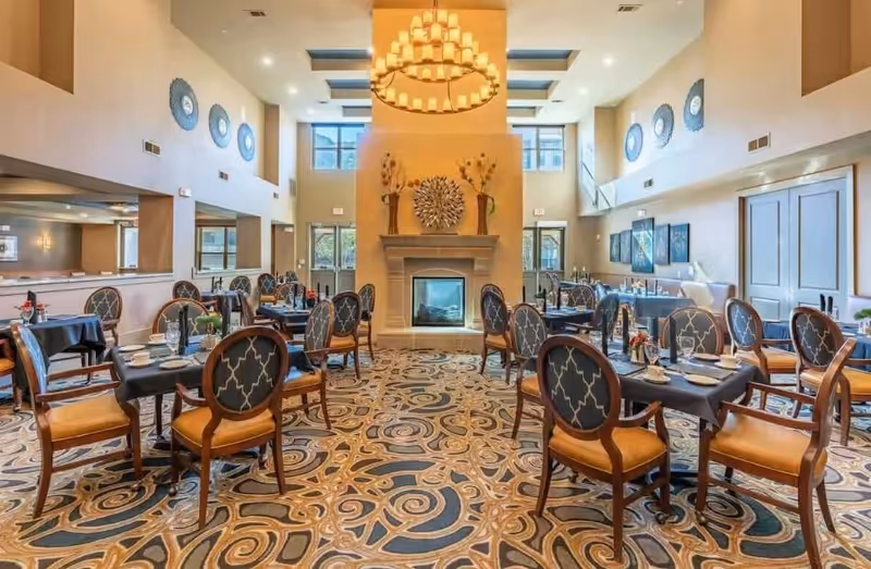 A spacious dining room with multiple tables covered in dark tablecloths, each set with plates, cups, and utensils. The room features elegant wooden chairs with patterned upholstery, a large decorative chandelier hanging from the ceiling, a fireplace centered on the far wall with decorative vases and artwork above it, and large windows allowing natural light to fill the space. The floor has a patterned carpet with swirling designs.