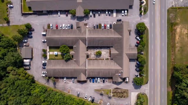 Aerial view of Compass Post Acute Rehabilitation facility showing the building complex with multiple wings, surrounding parking lots with parked cars, adjacent roads, and green landscaped areas.