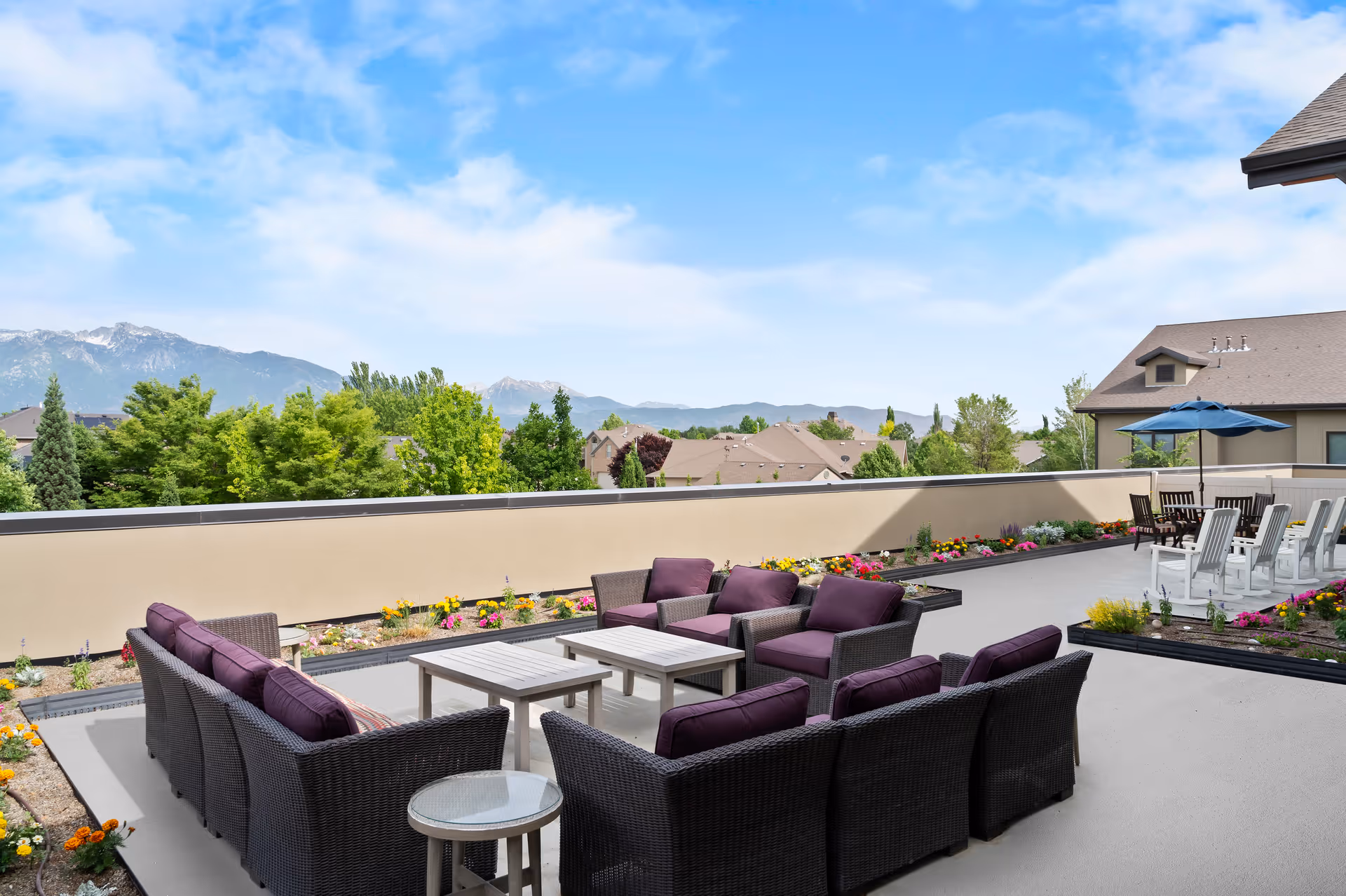 Rooftop patio with wicker seating and purple cushions, tables and flower beds overlooking houses and distant mountains.