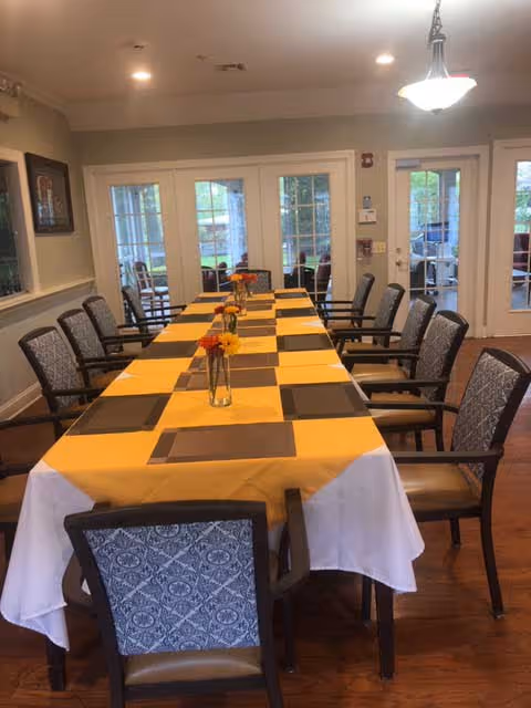A long dining table covered with a yellow and white tablecloth, set with black placemats and small vases holding orange and yellow flowers. The table is surrounded by cushioned chairs with patterned backs. The room has wooden flooring, beige walls, and large glass doors leading to an outdoor area.