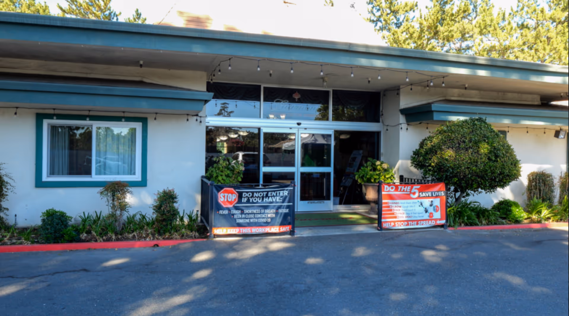 Exterior front entrance of a rehabilitation hospital with glass double doors, safety signage and potted plants.
