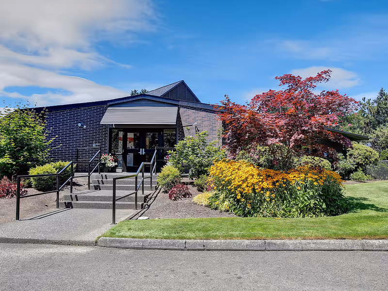 Front entrance of a brick building with steps, a wheelchair ramp and colorful landscaped flower beds under a blue sky.