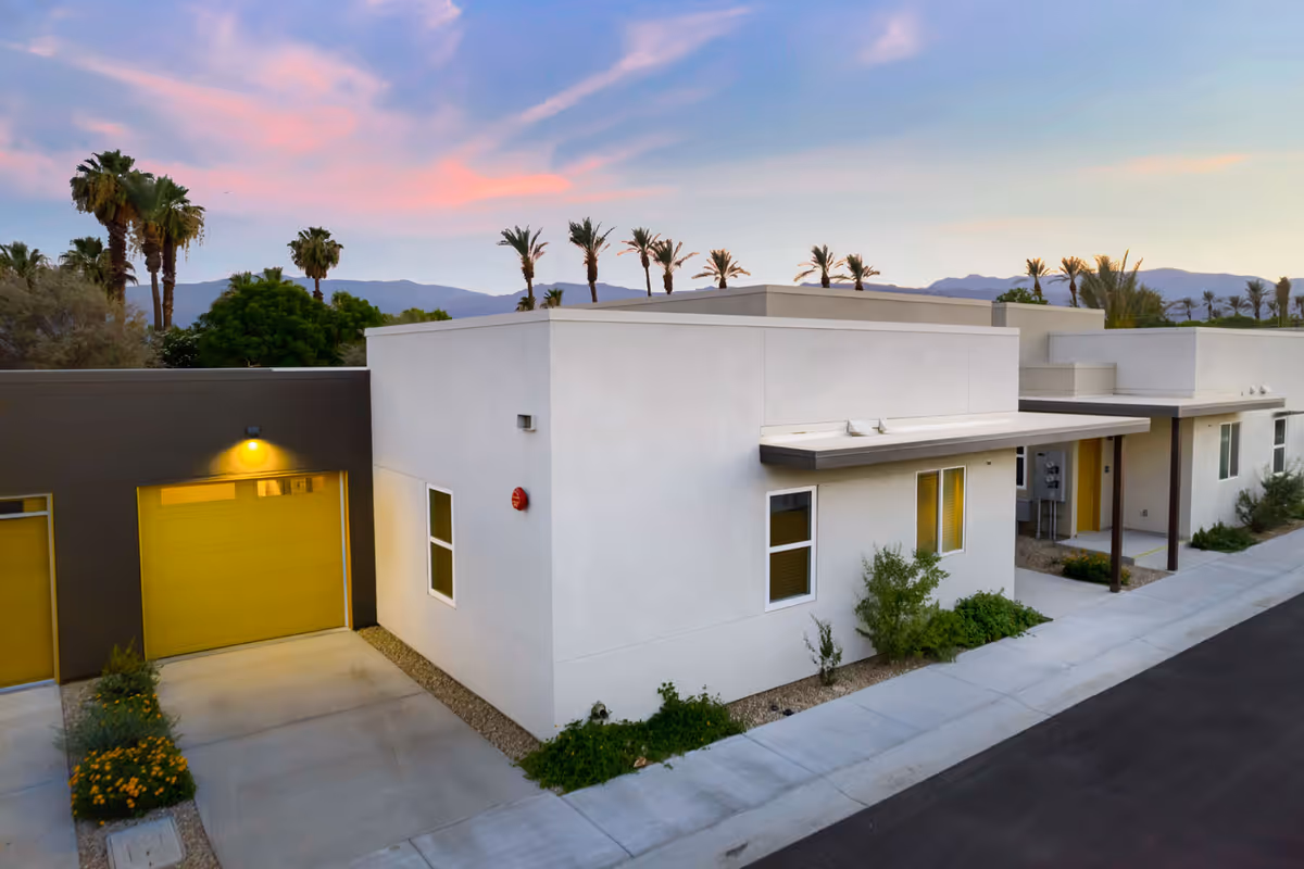Modern single-story white building with yellow garage doors and a driveway, palm trees and mountains under a colorful sunset sky.