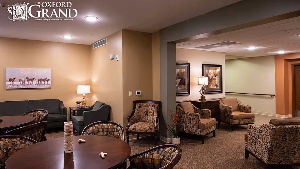 A cozy senior living community lounge area with multiple seating options including patterned armchairs and a dark green sofa. There are round tables with chairs around them, one table has a Jenga game set up. The walls are painted in warm tones and decorated with framed artwork and a lamp on a side table. The lighting is soft and inviting.