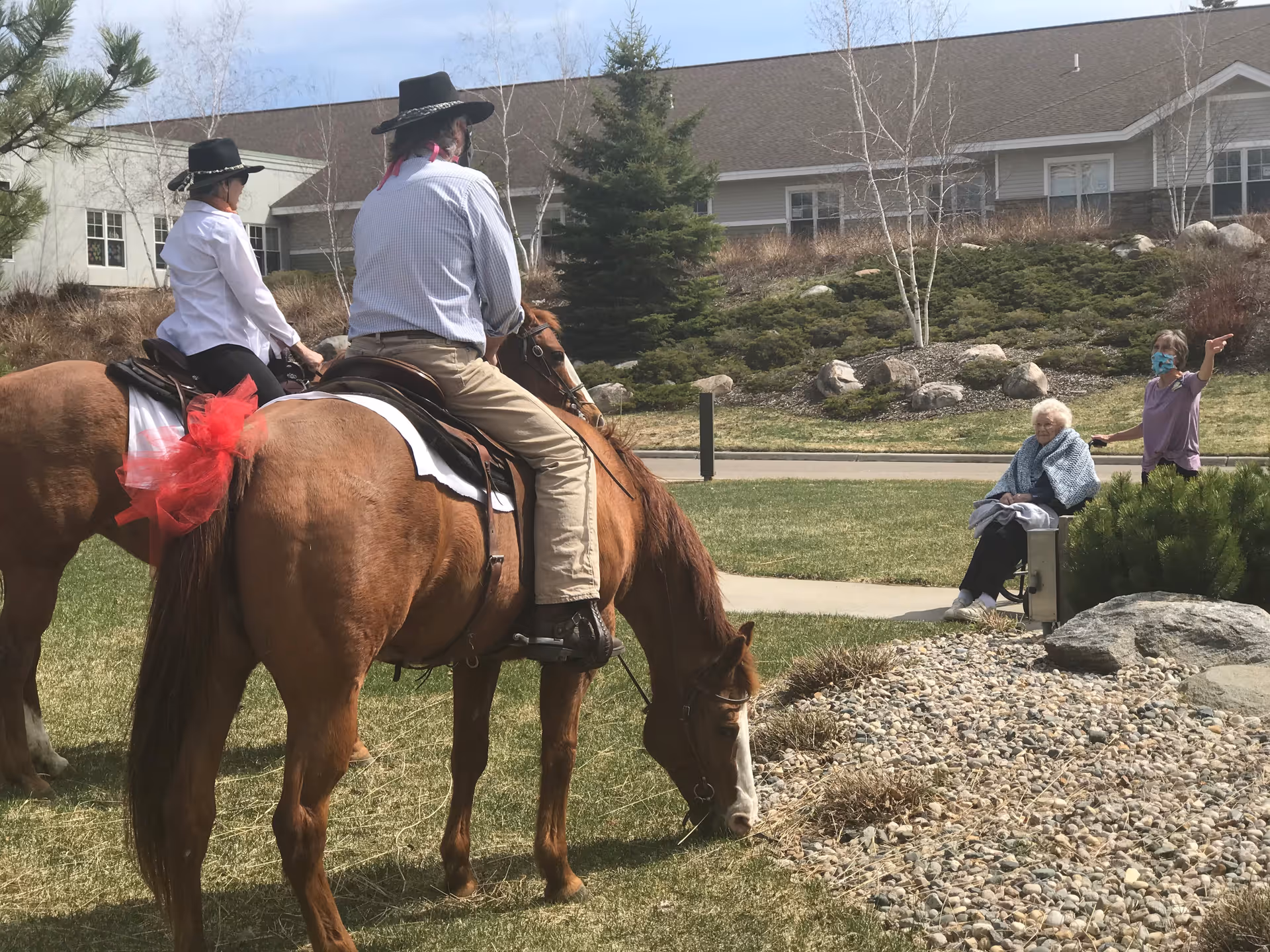 Two people wearing cowboy hats riding horses on a grassy area near a senior living facility. An elderly woman is sitting on a bench watching them, while another person wearing a mask is standing nearby pointing towards the riders. The background shows a building with windows and some landscaping with rocks and bushes.