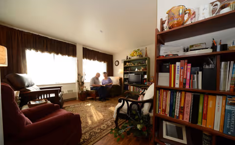 A cozy living room with two people sitting on a couch near a window with brown curtains. The room has a patterned rug, armchairs, a wooden bookshelf filled with books and decorative items, and a small TV on a stand.