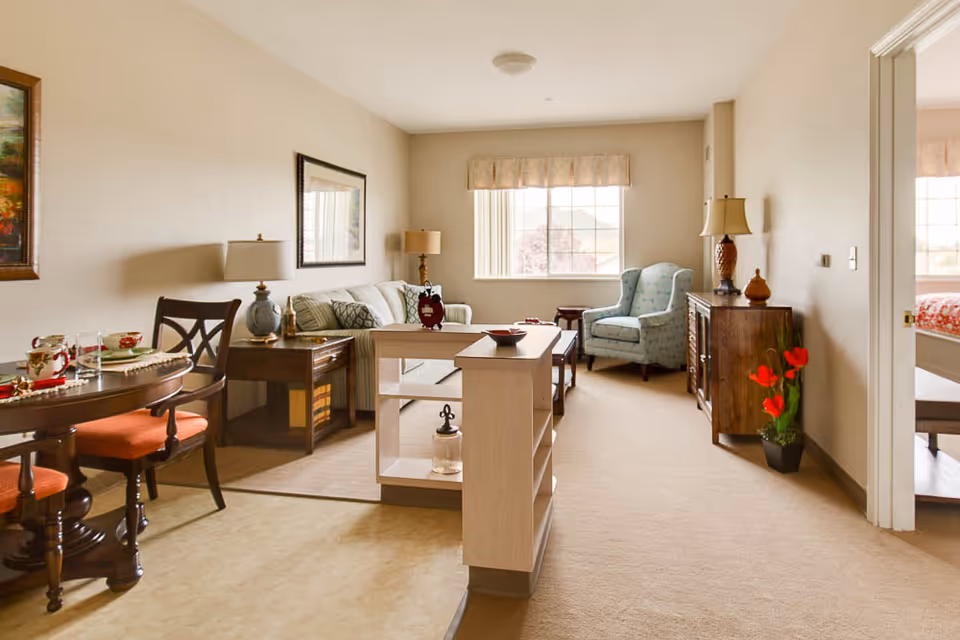 A bright and cozy living room area in a senior living facility with a beige carpet and walls. The room features a striped sofa with cushions, a wooden side table with a lamp, a blue upholstered armchair near a window with a valance, and a wooden cabinet with a lamp and decorative items. To the left, there is a round dining table with four chairs that have orange cushions, set with plates and glasses. A small shelving unit separates the living and dining areas. A doorway on the right leads to a bedroom with a bed visible.