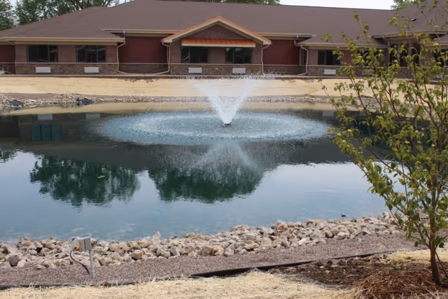 A pond with a water fountain in the center, surrounded by rocks and landscaping, with a single-story building in the background and a small tree on the right side.