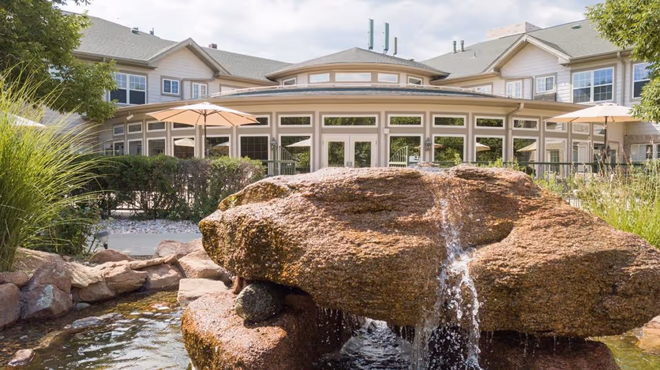 Outdoor view of a senior living facility named Gardens at Columbine featuring a large rock water fountain in the foreground, surrounded by greenery and plants, with a building and patio umbrellas visible in the background under a partly cloudy sky.
