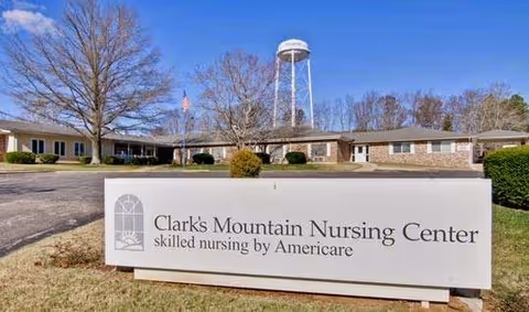 Exterior view of Clark's Mountain Nursing Center building with a large sign in the foreground displaying the facility's name and description. A water tower and leafless trees are visible in the background under a clear blue sky.