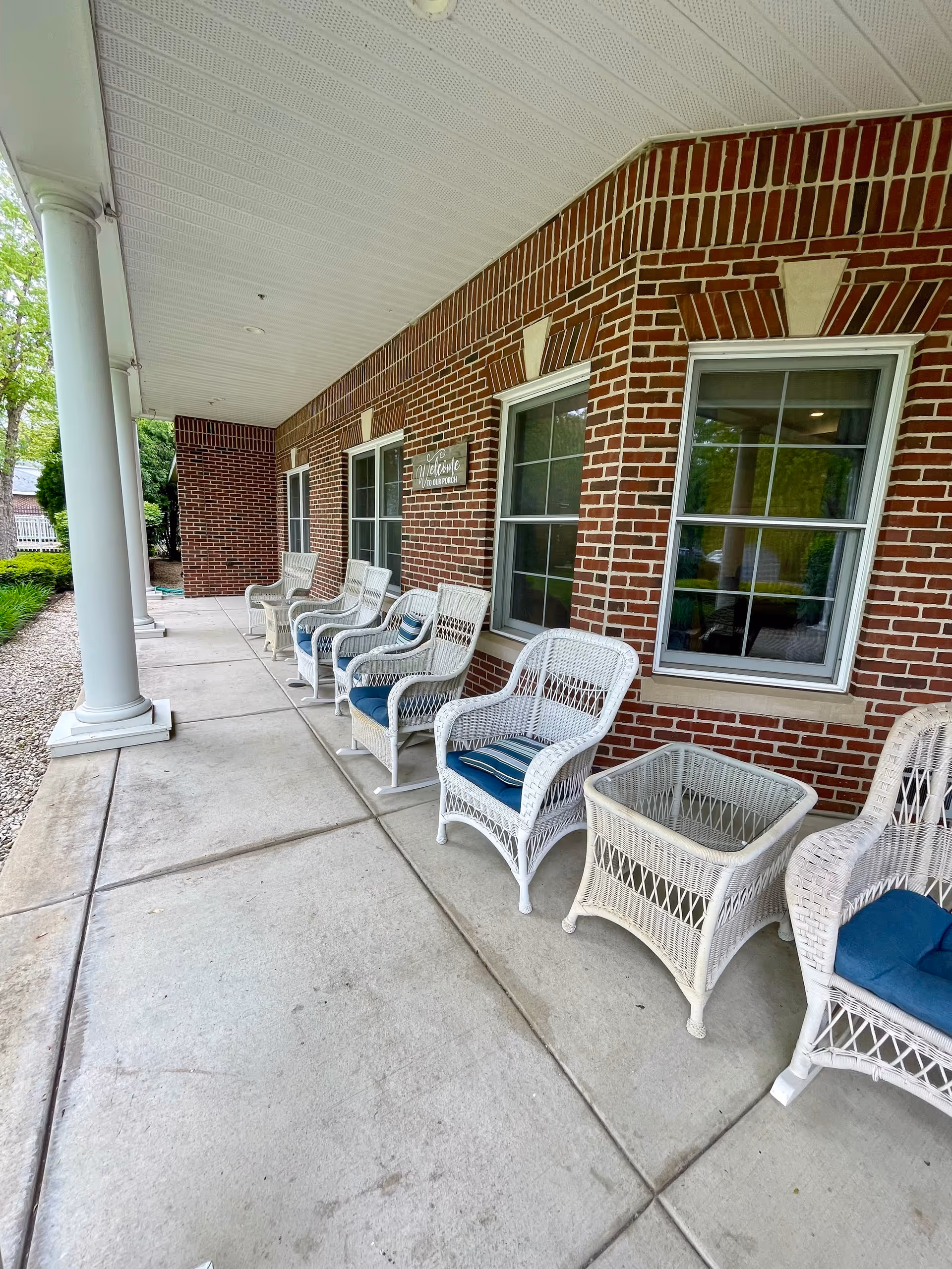 Covered outdoor porch area with white wicker chairs and a matching wicker table with blue cushions, set against a red brick wall with windows and a sign that reads 'Welcome to our porch'.