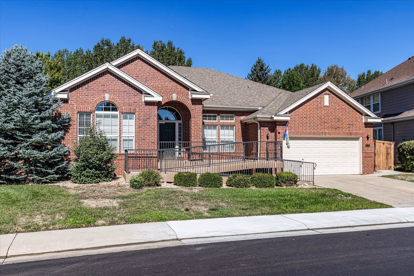 Front exterior view of a single-story brick house with a wheelchair accessible ramp leading to the front door, a two-car garage, and well-maintained landscaping including bushes and a large evergreen tree under a clear blue sky.