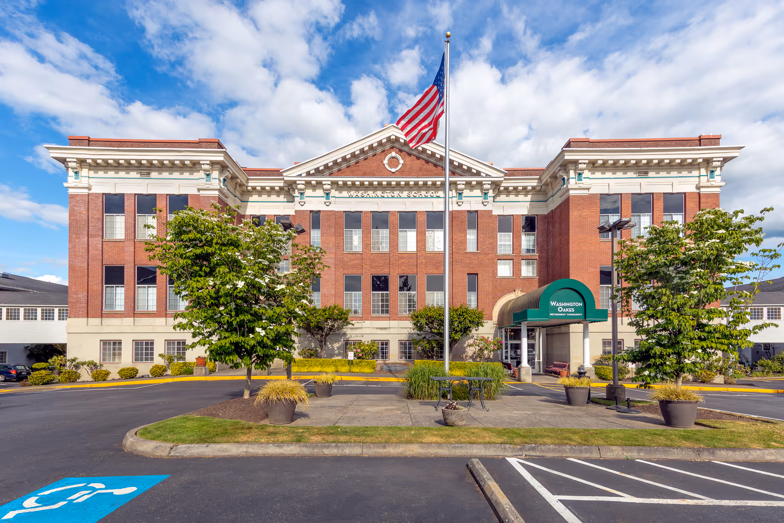 Front exterior view of a large brick building with white trim and multiple windows under a partly cloudy sky. An American flag flies on a flagpole in front of the building. There are trees, potted plants, and a small seating area near the entrance, which has a green awning labeled 'Washington Oakes Retirement Community'.