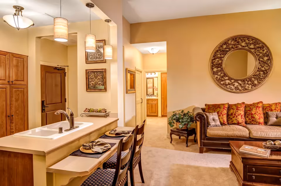 Interior view of a living space in Clermont Park Assisted Living featuring a kitchen counter with a sink and three chairs set with plates and napkins. The living room area has a brown leather sofa with patterned cushions, a wooden coffee table, a decorative round mirror on the wall, and a small side table with a plant. Warm lighting fixtures hang from the ceiling, and a hallway leading to another room is visible in the background.