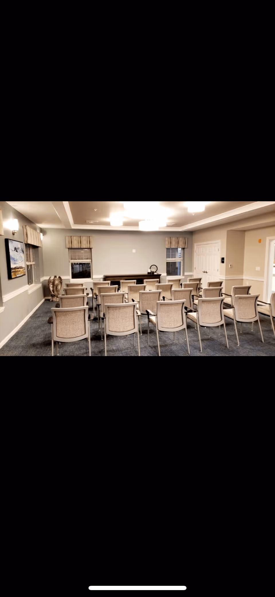 A meeting/activity room with rows of chairs facing a table and lectern in a senior living facility.