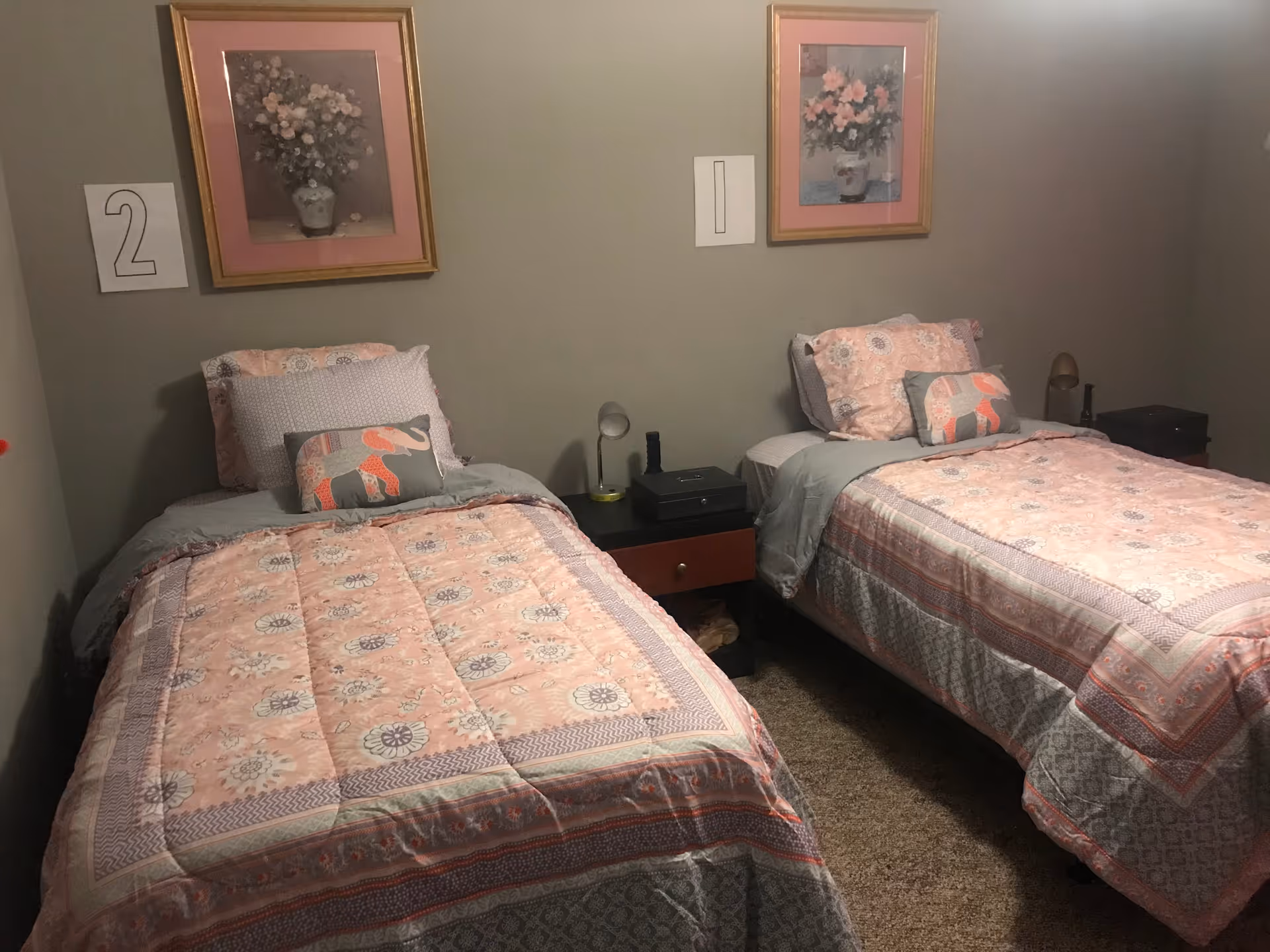 Small bedroom with two twin beds dressed in pink floral bedding, a nightstand between them and framed floral prints on the wall.