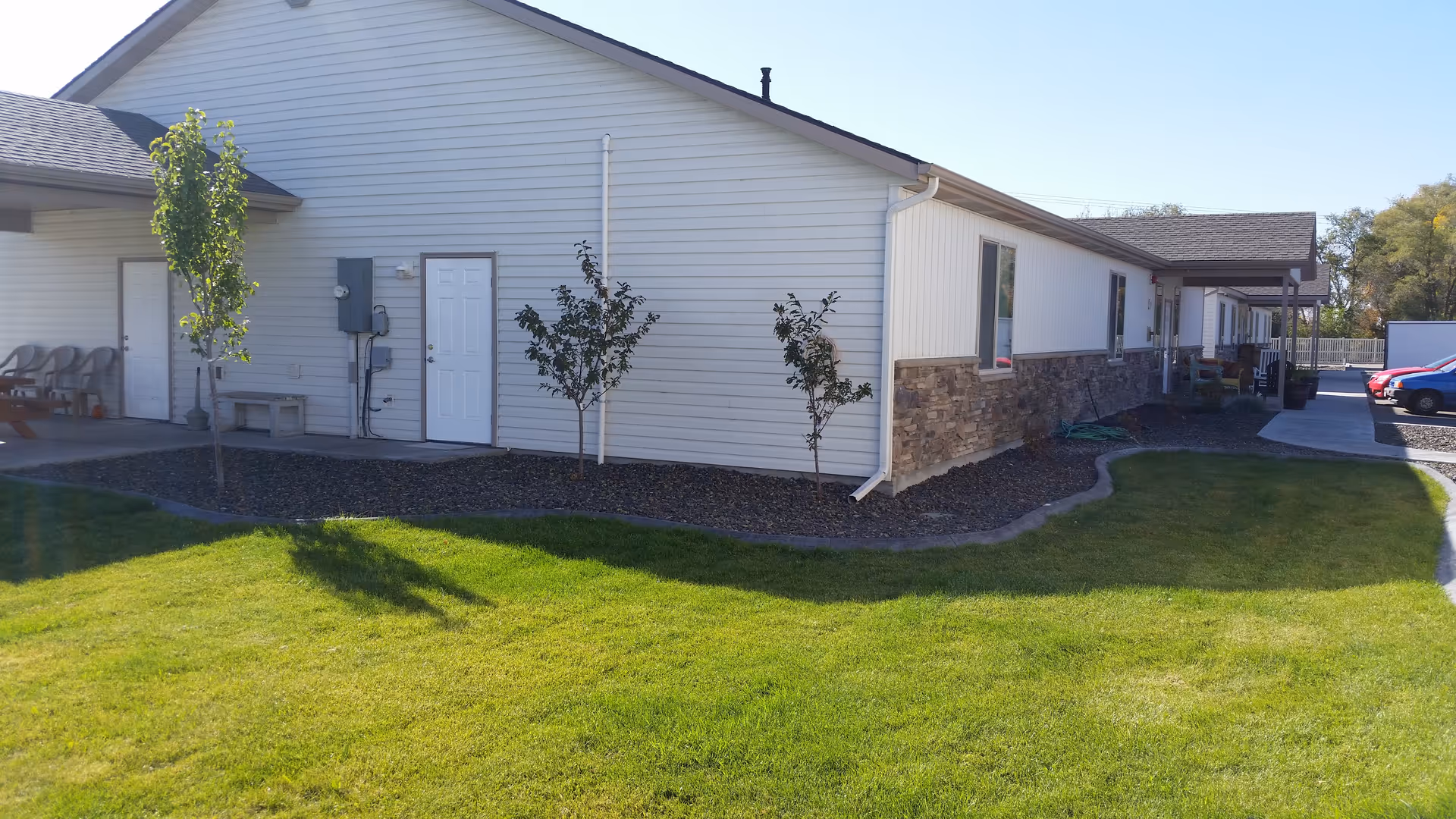 Exterior view of a single-story building with white siding and stone accents near the bottom. There are small trees planted along the side of the building, a green lawn in the foreground, and a paved parking area with cars visible in the background.