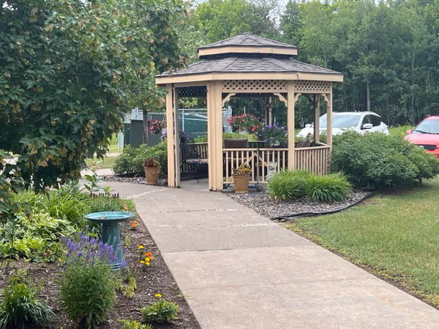 A wooden gazebo with a shingled roof is situated at the end of a paved walkway surrounded by green bushes, flowering plants, and a birdbath. There are hanging flower pots inside the gazebo, and parked cars are visible in the background near a wooded area.