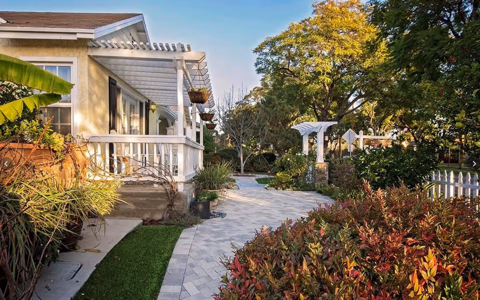 Front exterior of a cottage with a white porch, paved walkway, and landscaped garden under trees.