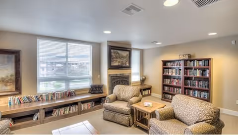 A cozy living room area in an assisted living facility featuring two patterned armchairs, a wooden coffee table, a fireplace with a framed painting above it, a large window with blinds, and bookshelves filled with books along the walls.