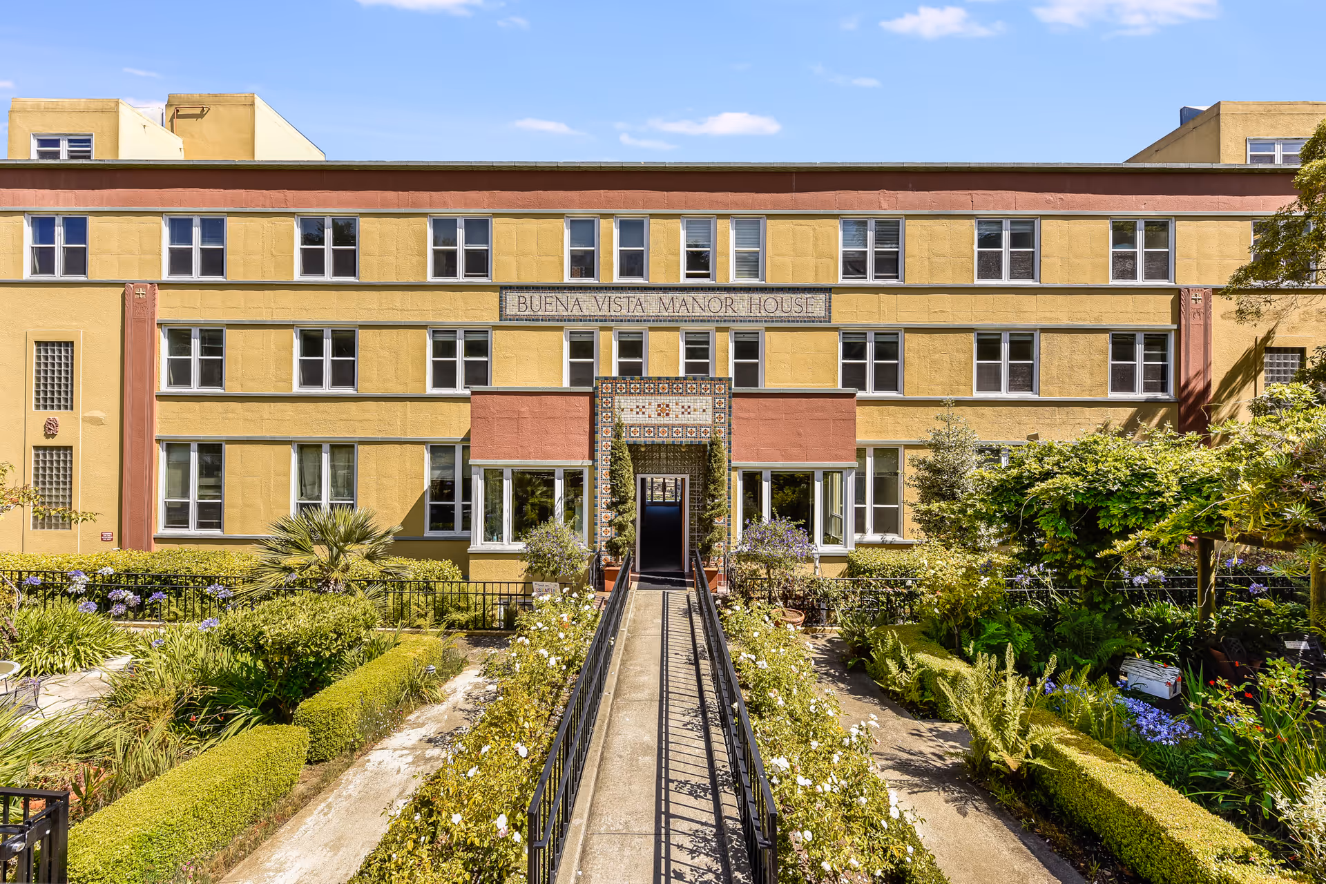 Front exterior view of Buena Vista Manor House, a three-story yellow building with multiple windows and a central entrance adorned with decorative tiles. A walkway with railings leads through a well-maintained garden with various plants and flowers towards the entrance under a clear blue sky.