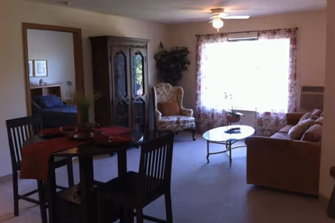 A cozy living area with a brown sofa, a glass coffee table, an upholstered armchair, and a tall wooden cabinet. A round dining table with four chairs is set with red placemats and a centerpiece. A large window with sheer floral curtains lets in natural light. A doorway leads to a bedroom with a bed and framed pictures on the wall.