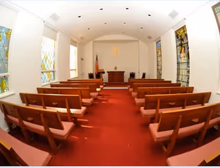 Interior view of a small chapel or worship room with wooden pews arranged in rows on a red carpet. At the front is an altar with a cross on the wall behind it, flanked by stained glass windows and an American flag to the left.