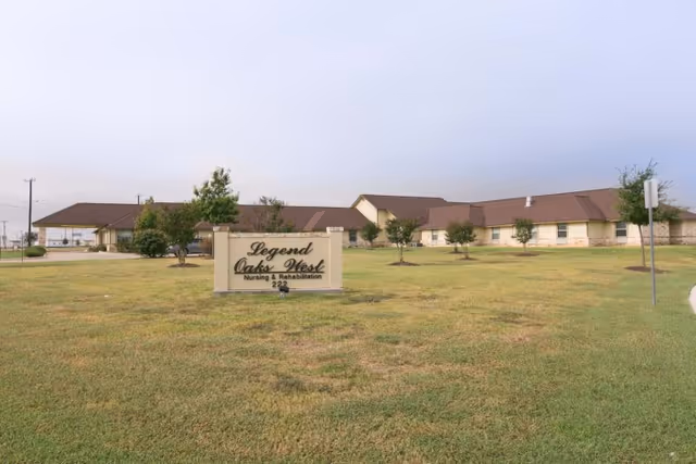 Exterior view of Legend Oaks West Nursing & Rehabilitation facility with a large grassy lawn and a sign displaying the facility name in front. The building has a beige exterior with brown roofing and several small trees planted around the lawn.