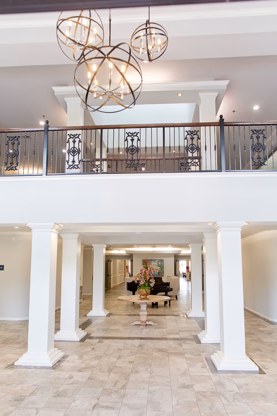 Spacious and elegant interior lobby area with white columns, tiled floor, a round wooden table with a flower arrangement in the center, and a black grand piano in the background. The upper level features a decorative wrought iron railing and modern spherical chandeliers hanging from the ceiling.